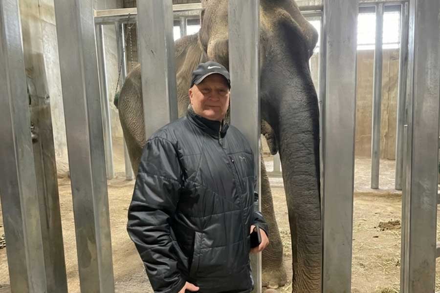 A man in a black jacket and cap stands smiling in front of an elephant behind vertical metal bars.