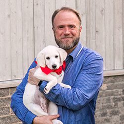 Dan Clevenger A bearded man in a blue shirt holds a white puppy with a red bandana, against a rustic wooden background.