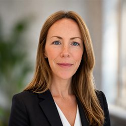 Katie Brennan Kane A woman with long brown hair, wearing a black blazer and white blouse, poses confidently. The background is softly blurred with a hint of greenery.