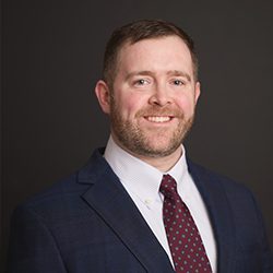 Kevin Mercer A man in a blue suit and maroon tie smiles warmly against a dark background. He has short hair and a neatly trimmed beard.