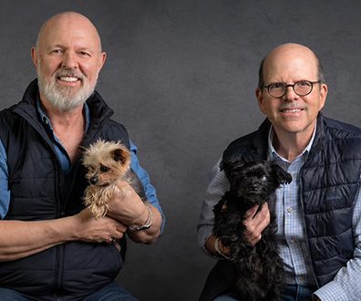 Two men sitting on a dark background, each with a dog beside them, creating a relaxed and friendly atmosphere.